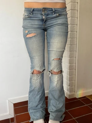Ljusblå slitna bootcut jeans - Säljer ett par ljusblå bootcut jeans med slitningar och hål vid knäna och låret. Jeansen har låg midja, fem fickor och knappgylf. Perfekta för en avslappnad och trendig look. Skriv ifall frågor. Har tecken på användning som visas på sista bild