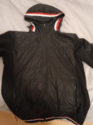 Moncler cardigan  - Teka inte o skriva! Snygg svart vindjacka från Moncler med huva och dragkedja framtill. Jackan har vita och röda detaljer på huvan och nederkanten samt Moncler-logga på ärmen. Praktiska fickor med dragkedja och sportig look. Storlek S (passar M) | BUD