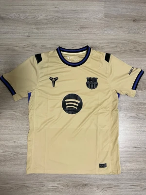 Barcelona bortatröja 25/26  - Snygg beige Barcelona-tröja från Nike med svarta detaljer och blåsvarta ränder vid krage och ärmslut. Tröjan har Dri-Fit-material som andas, korta ärmar och tryck med Spotify-logga på bröstet. Perfekt för dig som gillar sportig stil och fotboll.