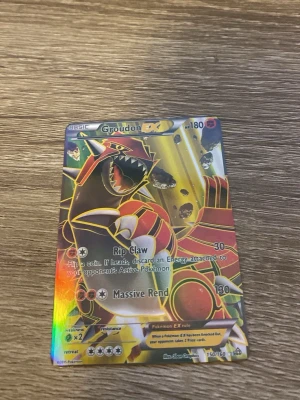 Groudon EX Pokémonkort - Säljer ett Groudon EX samlarkort från Pokémon med holografisk yta. Kortet har färgstarka illustrationer i rött, gult och svart och visar Groudon i en kraftfull pose. Perfekt för dig som samlar eller spelar Pokémonkort.
