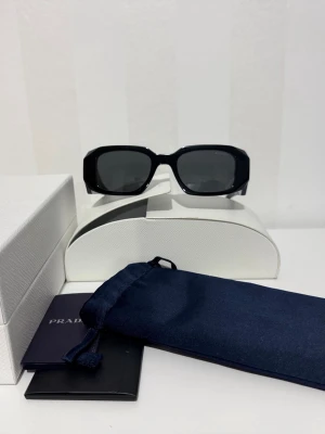Svarta solglasögon från Prada - Snygga svarta solglasögon från Prada med rektangulära bågar och mörka glas. Kommer med originalfodral, förvaringspåse och certifikat. Perfekta för dig som vill ha en stilren och exklusiv accessoar till sommaren.