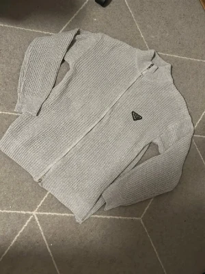Grå stickad ziptröja från Prada - Snygg grå stickad ziptröja från Prada med hög krage och ikonisk trekantslogga på bröstet. Tröjan har grovstickad struktur, långa ärmar och dragkedja framtill. Perfekt för lager-på-lager och en clean look.