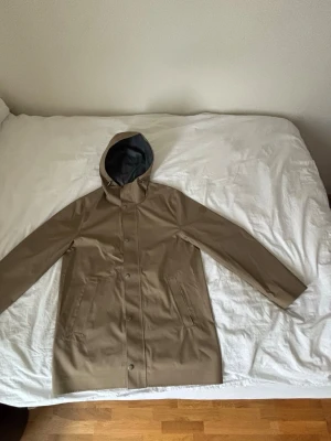 Beige regnjacka från GANT med huva - Stilren beige regnjacka från GANT i storlek M. Jackan har huva, två stora fickor framtill och stängs med tryckknappar. Materialet är vattenavvisande och insidan är fodrad i grått tyg. Perfekt för regniga dagar. (Orginalpris 4500)