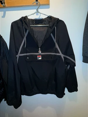 Svart vindjacka från Fila med huva - Snygg svart vindjacka från Fila med grå detaljer och halv dragkedja framtill. Jackan har en stor ficka på magen med knappstängning. Storlek M, passar även S. -Pris 299kr 