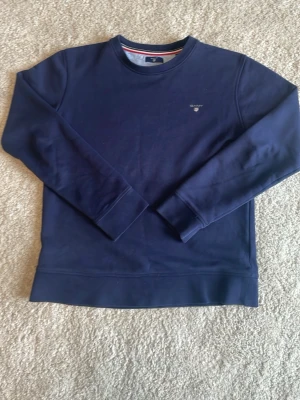 Marinblå sweatshirt från GANT - Klassisk marinblå sweatshirt från GANT med rund halsringning och diskret logga broderad på bröstet. Tröjan har långa ärmar och ribbade muddar vid ärmslut och nederkant. Perfekt för en avslappnad och stilren look.