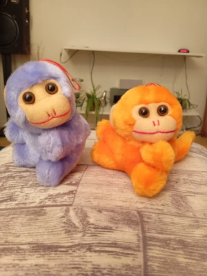 Två mjuka apor i plysch - Säljer två gulliga mjukisdjur i form av apor, en lila och en orange. Båda har stora bruna ögon, beige ansikte och röd detalj på huvudet för upphängning. Perfekta som dekoration eller present till någon som gillar söta gosedjur.