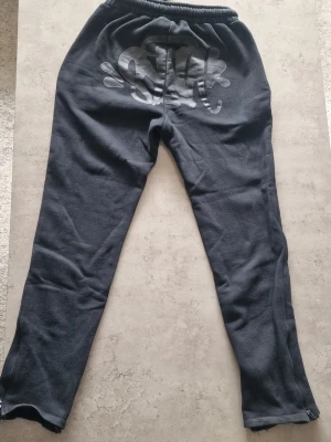 Svarta syna world mjukisbrall, Central cee - Svarta mjukisbyxor från syna world med resår i midjan och grov dragsko. Byxorna har fickor, dragkedjor vid bensluten och tryckt logga bak. Tillverkade i mjukt, fleeceliknande material för maximal komfort.