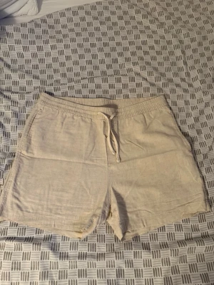 Beige linneshorts med snörning - Säljer ett par ljusbeige shorts i linne med avslappnad passform och snörning i midjan. Perfekta för varma dagar och har en enkel, stilren look. Shortsen har sidofickor och elastisk midja för extra komfort.