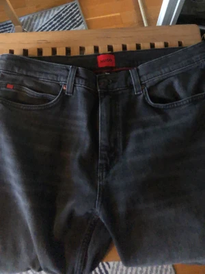 Svarta jeans från HUGO -  Helt oanvända Snygga svarta jeans från HUGO med klassisk femficksmodell och röda detaljer vid fickorna. Jeansen har en rak passform och är tillverkade i ett mjukt denimtyg. Perfekta för en stilren och modern look. Storlek 32/34