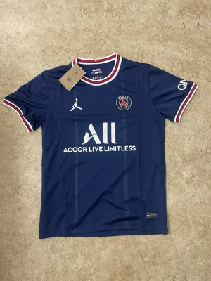 PSG matchtröja blå Jordan x Nike - Mörkblå PSG matchtröja med vita och röda detaljer på krage och ärmslut. Framtill finns PSG-logga, Jordan-logga och sponsortryck. Tillverkad i lätt och ventilerande material, perfekt för fotboll eller träning.