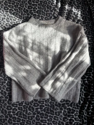 Grå ribbstickad tröja  - Mysig grå stickad tröja med ribbade vertikala ränder och avslappnad passform. Tröjan har långa ärmar och hög hals, perfekt för kyliga dagar. Enkel och stilren design som passar till det mesta.