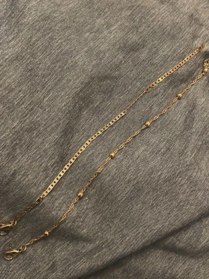 Två tunna guldarmband - Säljer två tunna armband i guld. Det ena har en enkel, platt kedja med rektangulära länkar och det andra har små runda pärlor längs en tunn kedja. Båda har smidiga karbinlås och passar perfekt för lager på lager-stil.