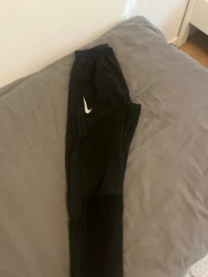 Svarta Nike dri-fit Aeroswift byxor - Säljer ett par svarta träningsbyxor från Nike med  reflex nike loggor  på båda sidor framtill. Byxorna har smal passform, resår i midjan och är tillverkade i ett lätt och stretchigt material som passar perfekt för sport och träning.