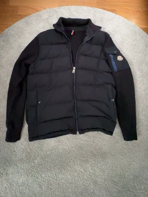 Svart pufferjacka från Moncler - Snygg svart pufferjacka från Moncler med quiltad front, stickade ärmar och avtagbar huva. Jackan har dragkedja framtill, två sidofickor med knapp och en ficka med Moncler-logga på ärmen. Perfekt för kalla vinterdagar och riktigt stilren.