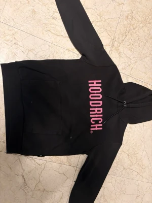 Svart HOODRICH hoodie med rosa tryck - Säljer en svart hoodie från HOODRICH med stort rosa tryck på bröstet. Hoodien har huva med dragsko och en stor magficka framtill. Tillverkad i mjukt bomullsmaterial som är skönt att bära. Perfekt för dig som gillar streetwear och vill sticka ut.