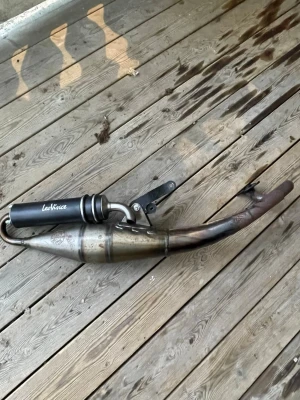 Svart avgassystem LeoVince scooter - Säljer ett svart avgassystem från LeoVince till scooter eller moped. Utrustad med ljuddämpare i svart metall och böjt rör i rostfritt stål. Medföljer fäste och packning. Perfekt för dig som vill ha bättre ljud och prestanda på din moppe.