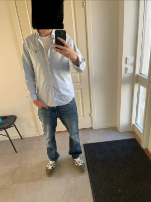 Ljusblå skjorta från Lyle & Scott, Storlek M - Säljer en stilren ljusblå skjorta från Lyle & Scott med klassisk button-down krage och bröstficka med den ikoniska gula örnloggan. Skjortan har långa ärmar och knäppning framtill. Tveka inte att skriva vid funderingar!!