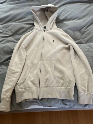 Beige hoodie från Polo Ralph Lauren - Snygg beige hoodie från Polo Ralph Lauren med dragkedja framtill och två stora fickor. Klassisk broderad logga på bröstet och mjukt material som känns skönt mot huden. Perfekt för en avslappnad och stilren look.