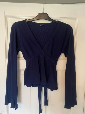 Mörkblå omlottopp med knytband - Snygg mörkblå omlottopp med långa ärmar och knytband i midjan. Djup v-ringning framtill och mjukt, stretchigt material som sitter bekvämt. Perfekt att styla med jeans eller kjol för en trendig look.