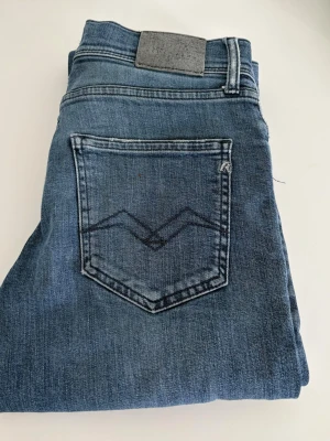 Blå jeans från Replay med fickdetalj - Tja, Säljer ett par klassiska blå jeans från Replay med snyggt broderad bakficka och dragkedjegylf. Jeansen har en rak passform och är tillverkade i mjukt denimtyg. Perfekta för en avslappnad och trendig look. Knappen som man knäpper har lossnat så det får man fixa 
