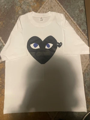 Vit t-shirt med svart hjärta print - Vit t-shirt med stort svart hjärta och ögon på bröstet samt ett mindre hjärta bredvid. T-shirten har rund hals och korta ärmar. Materialet känns som bomull och trycket ger en cool och unik vibe.