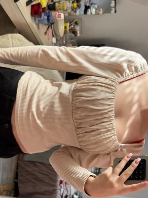 Beige ribbad offshoulder topp - Trendig beige offshoulder topp med ribbad struktur och lång ärm. Toppen har en snygg rynkad detalj över bysten och sitter tight för en smickrande passform. Perfekt för dig som vill ha en stilren men ändå unik look.
