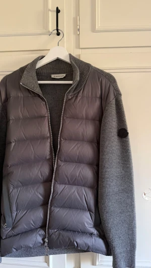 Grå Moncler cardigan  - Snygg grå dunväst från Moncler med stickade ärmar och krage. Västen har quiltad front i dunmaterial och dragkedja framtill. Ärmarna är ribbstickade i matchande grått och har en diskret Moncler-logga på vänster ärm. Perfekt för lager-på-lager och streetwear. Herr moncler cardigan, perfekt nu till denna tiden! Inga yttre skador eller liknande, super bra skick! 