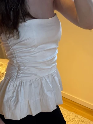 Vit axelbandslös peplum topp - Snygg vit axelbandslös topp med peplumvolang nedtill. Toppen har en bandeauformad överdel och är figurnära med lätt rynkade detaljer i midjan. Perfekt för dig som vill ha en stilren och trendig look.🤍🌟🪩😍🎉🌸