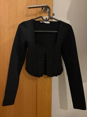 Svart croppad långärmad topp - Trendig svart croppad topp med lång ärm och fyrkantig urringning. Toppen har knäppning framtill och en figurnära passform som ger en snygg siluett. Perfekt att styla med höga jeans eller kjol.