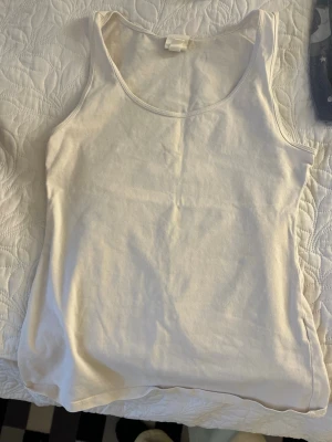 Vit enkel linnetopp - Säljer en basic vit linnetopp i mjuk bomull. Toppen är ärmlös med rund halsringning och har en klassisk, enkel design som passar till det mesta. Perfekt att styla lager på lager eller bära som den är.