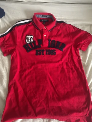 Röd pikétröja från Tommy Hilfiger - Snygg röd pikétröja från Tommy Hilfiger med broderad logga och text 'HILFIGER EST 1985' på bröstet. Tröjan har korta ärmar, krage med knappar och marinblå detaljer på axlarna och kragen. Perfekt för en sportig och avslappnad stil.