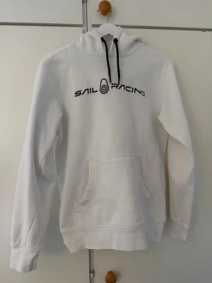 Vit hoodie från Sail Racing - Vit hoodie från Sail Racing med svart logga och text på bröstet. Har några fläckar på framsidan som säkert går bort för den som använder fläckborttagningsmedel och dylikt. Annars är den väldigt lite använd så i övrigt är det jättefint skick på den.