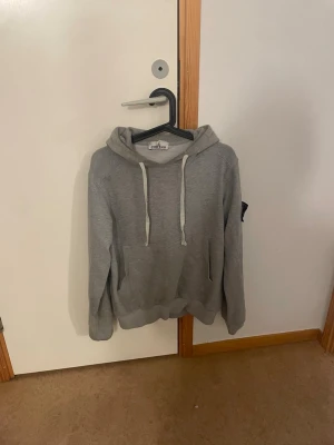 Grå hoodie från Stone Island - Säljer en stilren grå hoodie från Stone Island med klassisk huva och vita dragsnören. Hoodien har en stor magficka och den ikoniska Stone Island-loggan på vänster ärm. Perfekt för dig som gillar streetwear och enkel design.