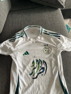 Algeriet fotbollströja Adidas vit - Säljer en vit Algeriet fotbollströja från Adidas med gröna och blå detaljer, emblem på bröstet och arabiskt tryck framtill. Tröjan har korta ärmar, rund hals och klassiska Adidas-ränder på axlarna. Perfekt för fotbollsträning eller match.