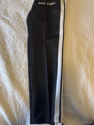 Palm Angels svarta mjukisbyxor - Palm Angels svarta mjukisbyxor med vita sidoränder och dragkedjor vid bensluten. Klassisk streetwear-look med logga framtill och baktill i vitt. Tillverkade i ett glansigt, sportigt material som ger en snygg och avslappnad vibe. Priset kan diskuteras lite