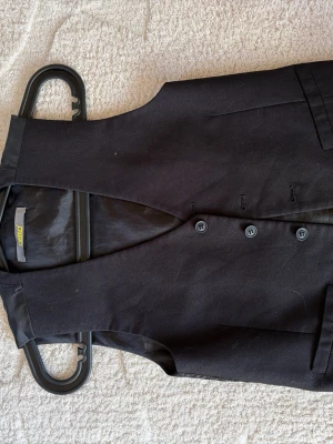 Svart väst från Demo med knappar - Klassisk svart väst från Demo med tre knappar framtill och enkel passform. Västen har en stilren look och är tillverkad i ett slätt tyg som ger en clean känsla. Perfekt att styla över en skjorta för en snygg och tidlös outfit.