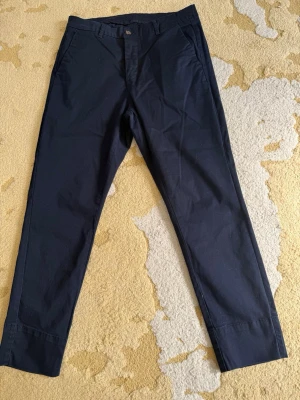 Mörkblå chinos Happy - Snygga mörkblå chinos med klassisk rak passform och diskreta fickor i sidorna. Byxorna har en stilren look och är tillverkade i ett mjukt bomullstyg som känns bekvämt hela dagen. Perfekta för dig som gillar en clean och enkel stil. 