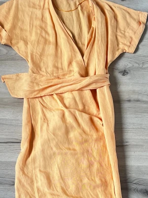 Persikofärgad wraparound klänning i bomull - Klänning i en mjuk persikofärg med korta ärmar och knytskärp i midjan. Tillverkad i tunt, luftigt bomullstyg som känns skönt mot huden. Perfekt för en sommar