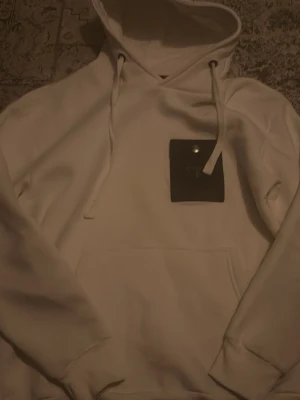 Vit hoodie från prada med ficka - Säljer en clean vit hoodie från Prada med en unik svart ficka på bröstet och den klassiska Prada-loggan i metall. Hoodien har huva med dragsko och en stor magficka. Perfekt för dig som gillar exklusiva streetwear-plagg.