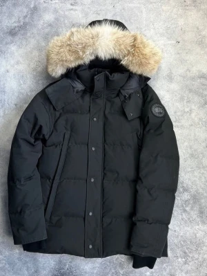 Canada goose black label - Säljer en svart parkasjacka från Canada Goose med avtagbar huva och lyxig pälskant i beige. Jackan har diskret logotyp på ärmen, dragkedja och knappar framtill samt två snedställda fickor. Perfekt för kalla dagar och riktigt snygg streetstil.