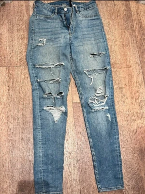 Blå ripped jeans från H&M Divided - Säljer ett par blå skinny jeans från H&M Divided med flera slitningar och hål på framsidan. Jeansen har hög midja och klassisk femficksmodell. Perfekta för en avslappnad och trendig look. Materialet är bomullsdenim med lite stretch.