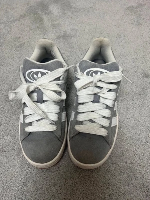 Gråa Adidas sneakers med vita detaljer - Snygga gråa Adidas sneakers med klassiska vita ränder på sidorna och breda vita skosnören. Ovandelen är i mockaliknande material och har en tydlig Adidas-logga på plösen. Perfekta för en avslappnad streetwear-look. Betalning sker via vanlig Swish! 