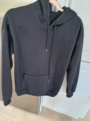 Svart hoodie  - Klassisk svart hoodie med huva och dragsnören. Hoodien har en magficka framtill och ribbade muddar vid ärmslut och nederkant. Tillverkad i mjukt, fleeceliknande material på insidan för extra komfort. O