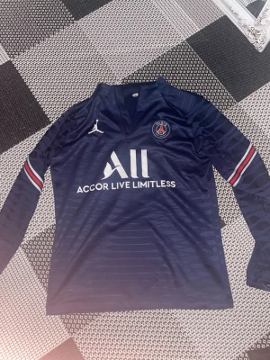 Mörkblå PSG långärmad matchtröja - Snygg mörkblå långärmad PSG matchtröja från samarbetet med Jordan. Tröjan har vita och röda detaljer på ärmarna, PSG-logga på bröstet och texten 'Accor Live Limitless' framtill. Tillverkad i ett lätt och ventilerande material, perfekt för fotboll eller träning.