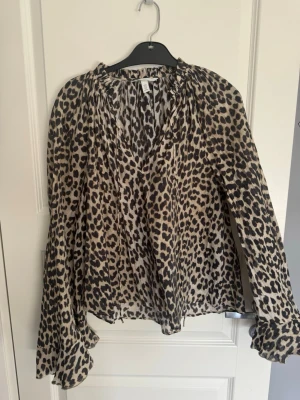 Leopardmönstrad blus från H&M - Snygg blus från H&M med leopardmönster i beige och svart. Blusen har v-ringning, långa vida ärmar och rynkade detaljer vid halsen. Tillverkad i ett tunt, luftigt material som ger en avslappnad och trendig look.