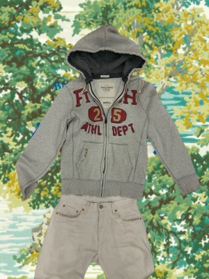  hoodie Abercrombie & Fitch - Snygg grå hoodie från Abercrombie & Fitch med dragkedja och huva. Stora röda bokstäver och siffran 25 framtill ger en sportig vibe. Mjuk insida och två fickor fram. Perfekt för en avslappnad streetstyle-look.