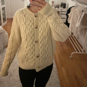 Beige stickad kofta med knappar - Mysig beige stickad kofta med flätmönster och knappar framtill. Perfekt för kyliga dagar med sin varma och bekväma design. Långärmad med ribbade muddar vid ärmslut och nederkant.