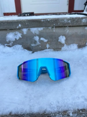 Blå sportiga solglasögon - Säljer ett par riktigt snygga sportiga solglasögon med spegelblå lins och kantig, futuristisk form. Perfekta för dig som vill sticka ut och få en cool look. Linsen har en blå och lila ton som reflekterar ljuset. Nya unisex