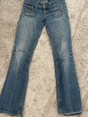 Blå bootcut jeans från ONLY - Snygga blå bootcut jeans från ONLY med slitningar och en trasig detalj på vänster ben. Klassisk femficksmodell med kontrastsömmar och knappgylf. Tillverkade i denim med en cool tvättad look. Säljes då de är för små!