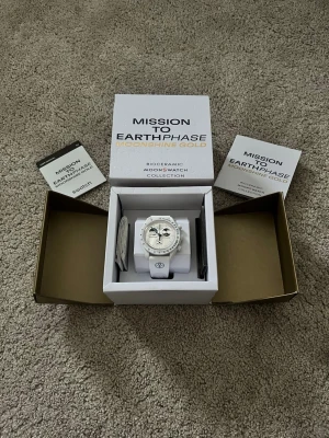 Omega x Swatch MoonSwatch Moonshine Gold - Säljer en vit Omega x Swatch Mission to Earthphase Moonshine Gold Snoopy från Bioceramic MoonSwatch Collection. Klockan har en rund urtavla med detaljer i svart och guld, samt vit textilrem. Snygg och unik design med månfaskomplication.